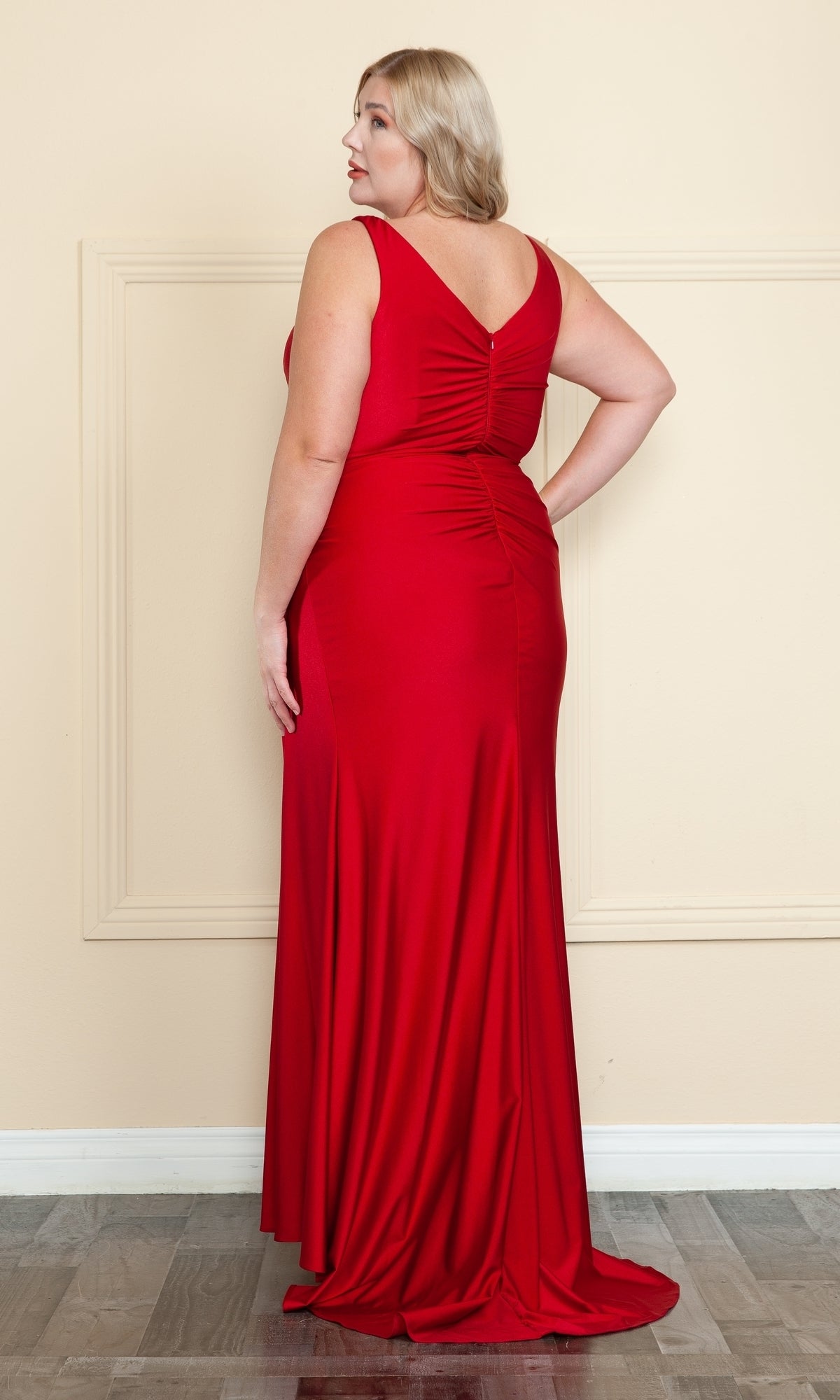 A woman stands on a wood floor, facing away, wearing the Faux-Wrap Plus-Size Long Prom Dress W1114—a sleeveless red gown with a V-shaped back, ruched waist, and gathered detailing.